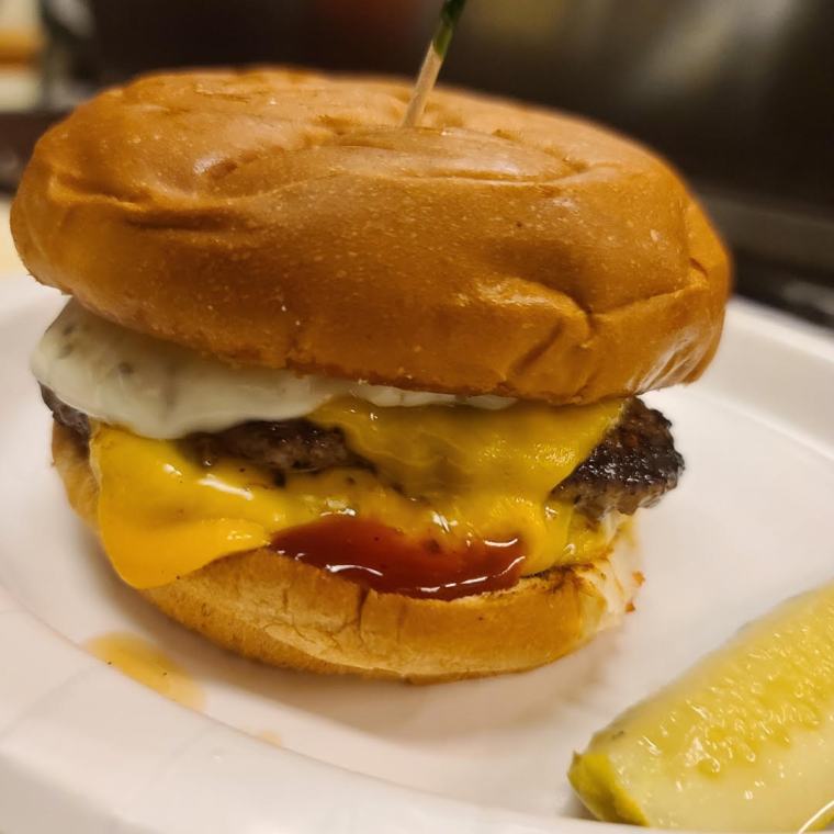 The Charlie Burger on a white plate — two patties with American cheese, BBQ and tartar sauce, a pickle spear alongside