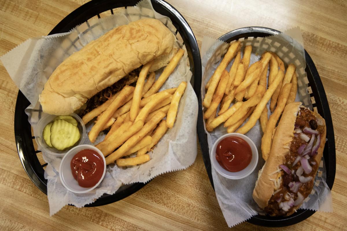 Two baskets on the butcher block from overhead — a steak sub and a chili dog, each with a pile of fries