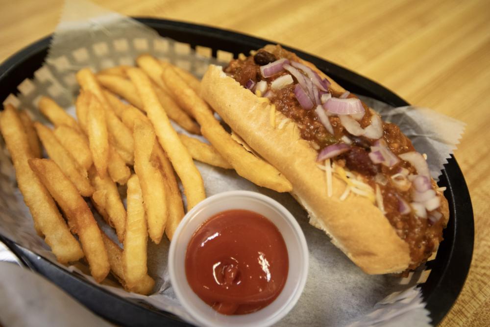 A chili dog under shredded cheese and red onion, fries and ketchup alongside