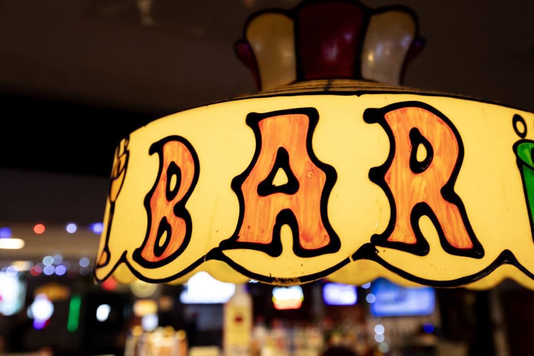 The stained-glass BAR lamp lit above the taps, the bar bottles glowing out of focus behind it