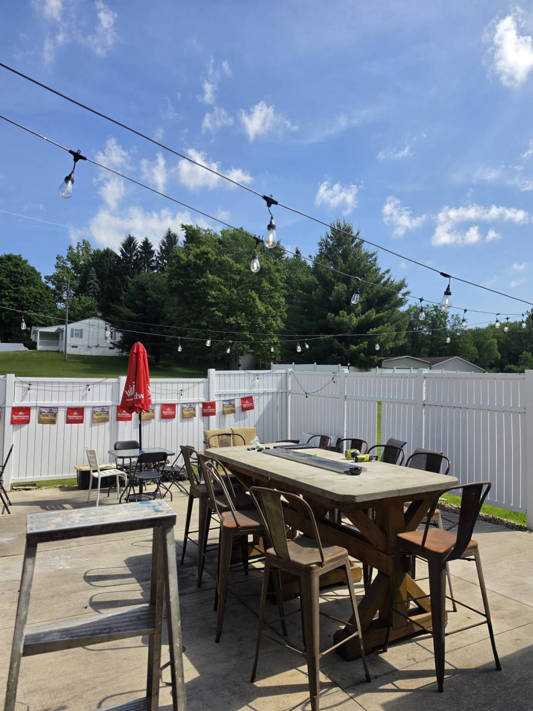 The patio in daylight — white fence, string lights, tall wood tables and a red umbrella