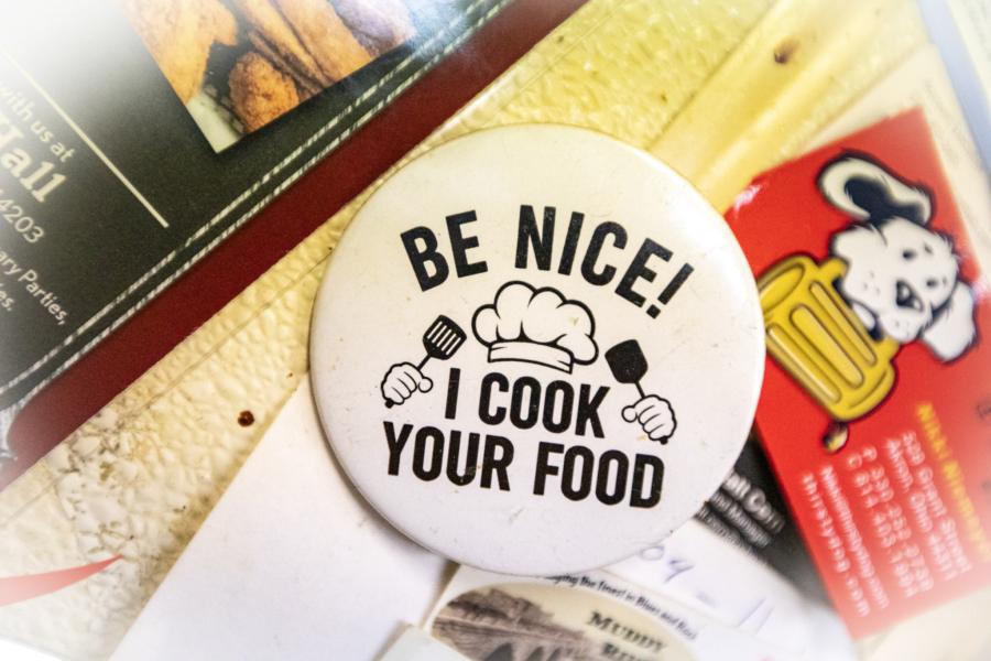 A pin badge sitting on a stack of menus reading "BE NICE! I COOK YOUR FOOD" around a chef's hat and two spatulas